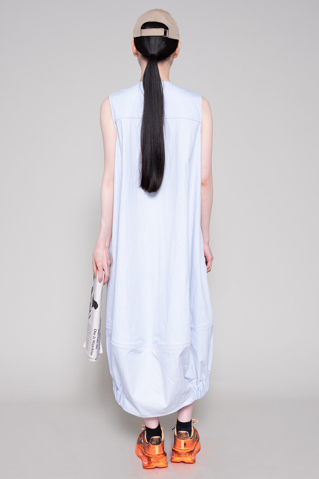 Person wearing a light blue sleeveless dress with beige cap and orange sneakers, holding a newspaper against a plain background.