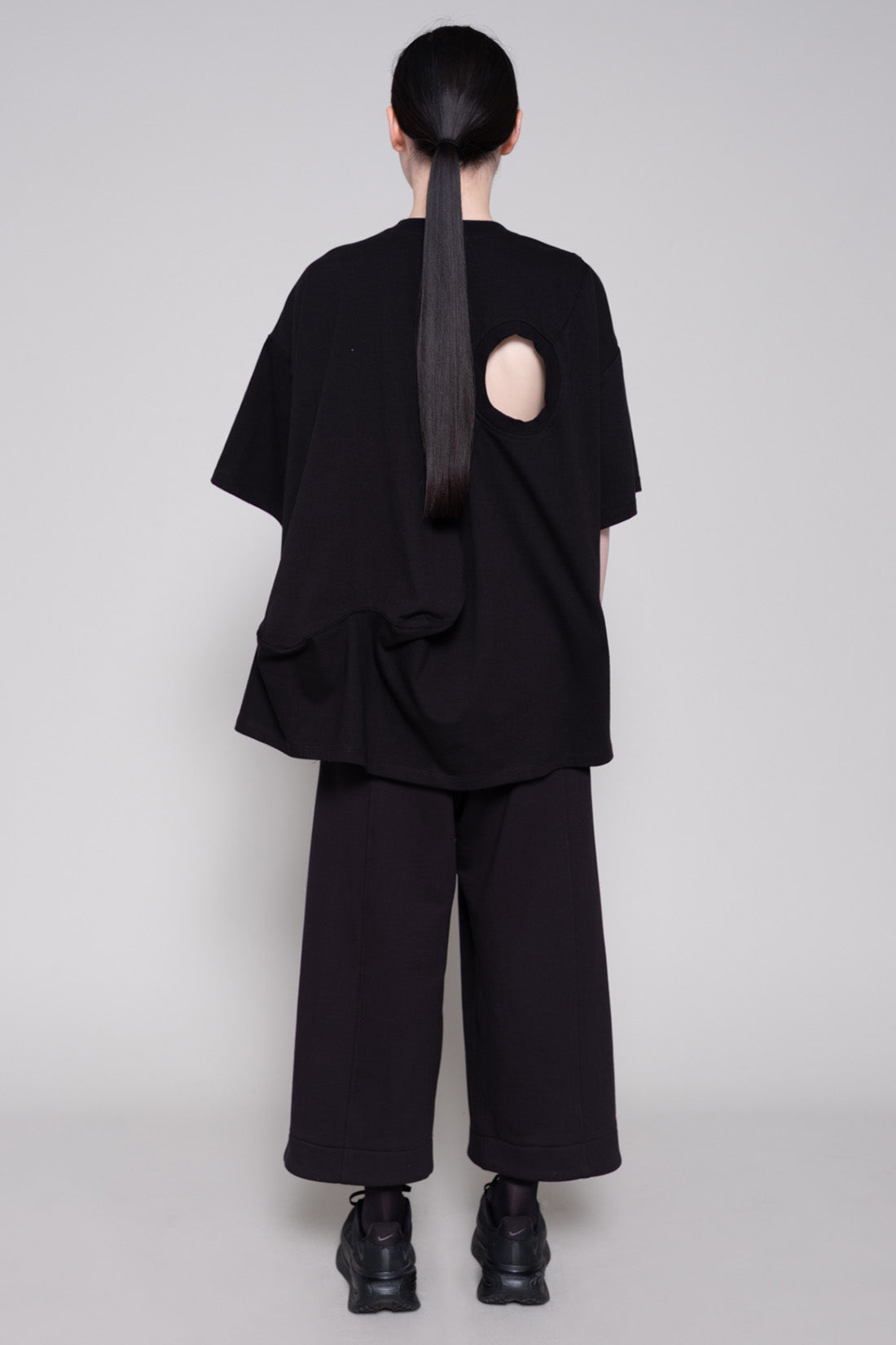 Person wearing a black tee with cutout and pants against plain background