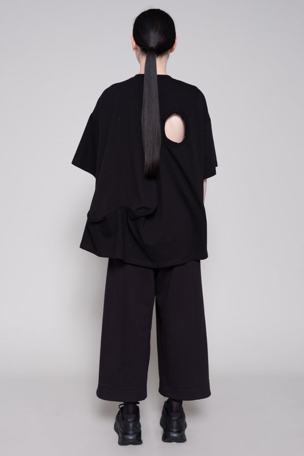 Person wearing a black tee with cutout and pants against plain background