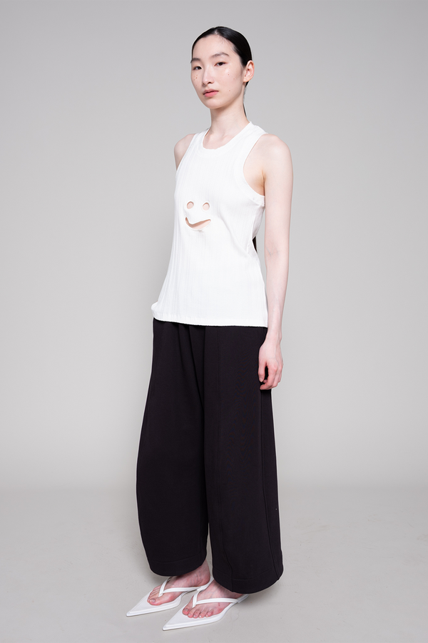 Person wearing a white sleeveless top with a smiley face design and black pants on a gray background