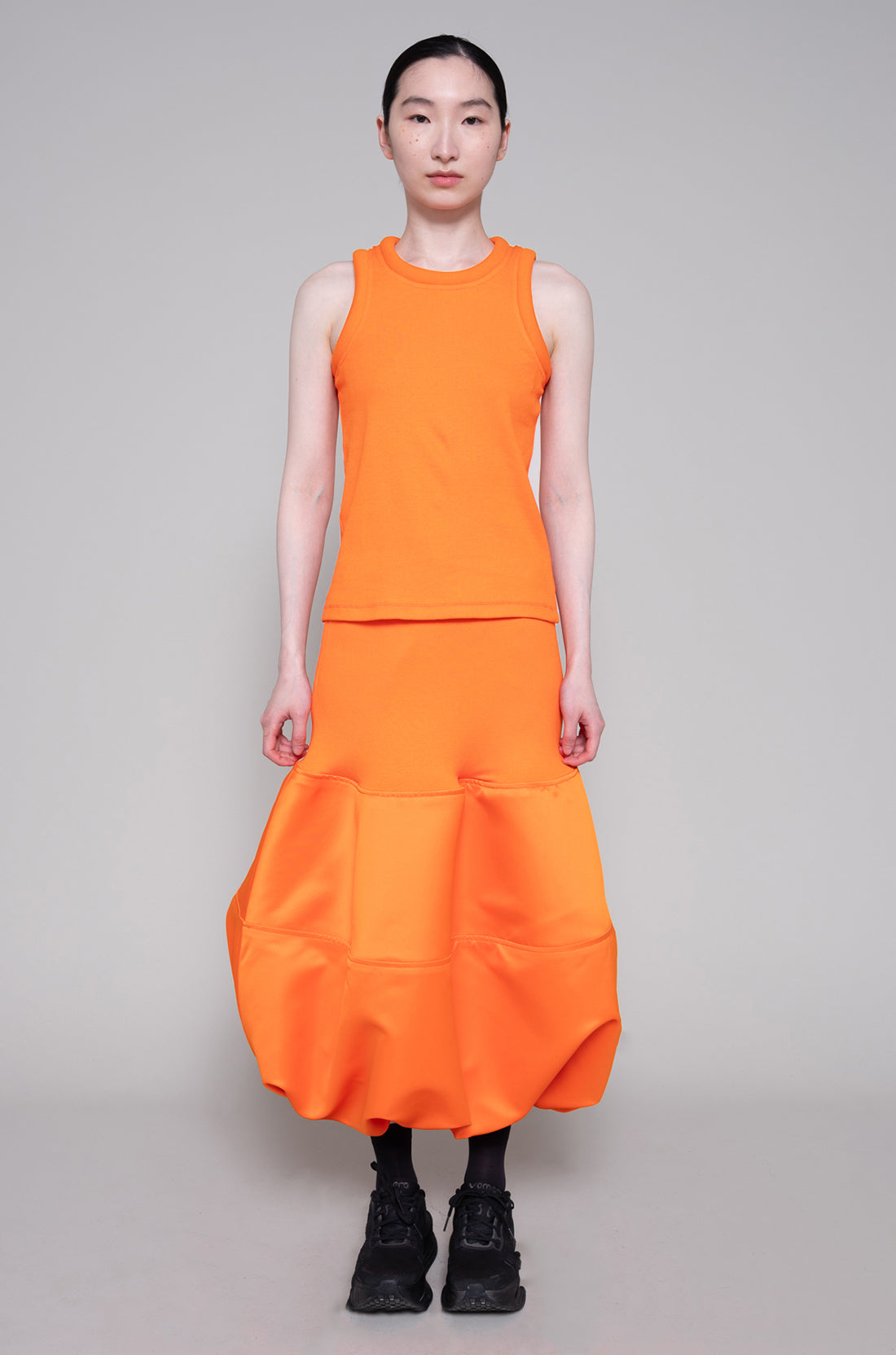 Person wearing sleeveless orange top and skirt with black sneakers on plain background