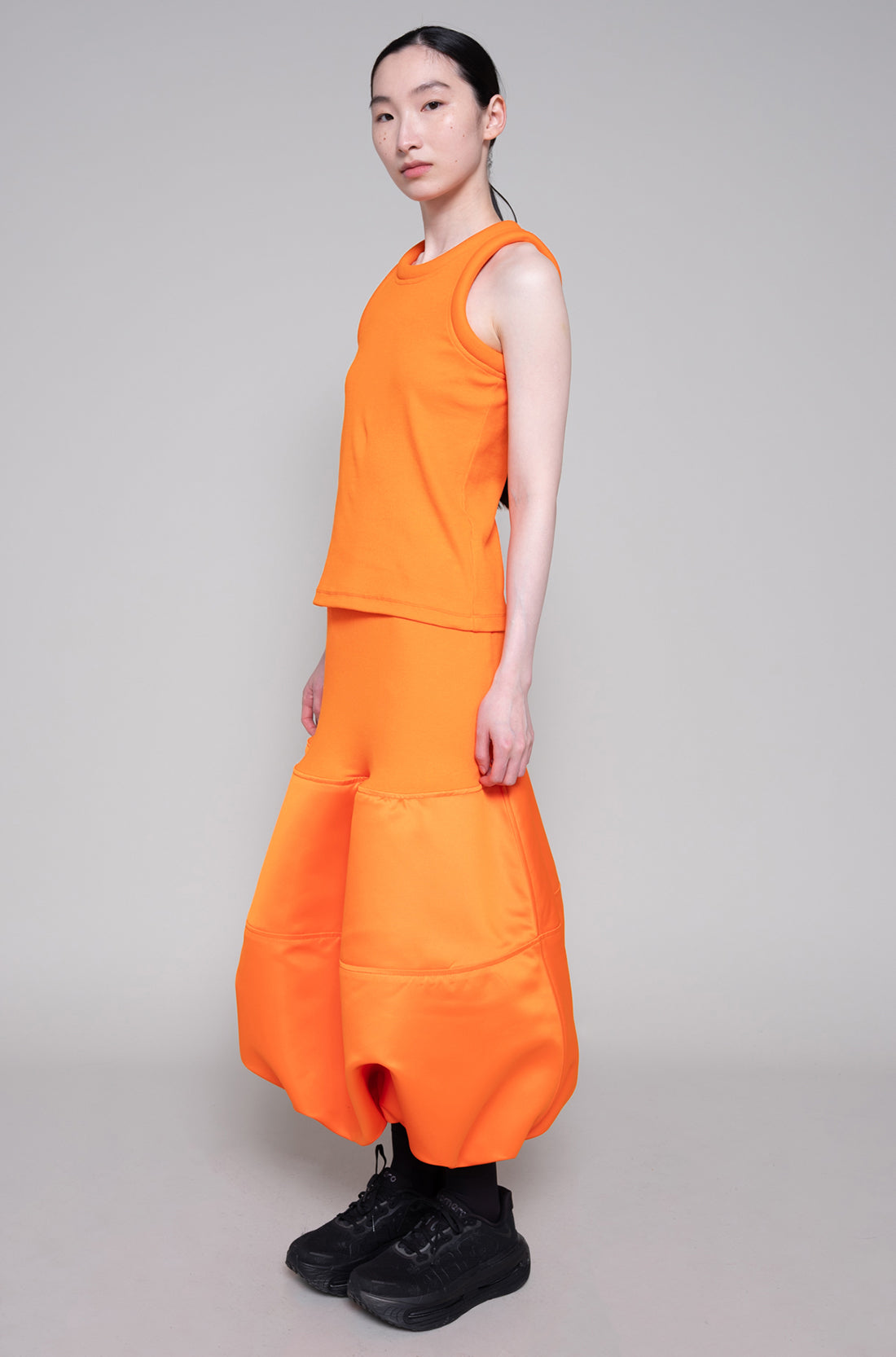 Person wearing sleeveless orange top and skirt with black sneakers on plain background