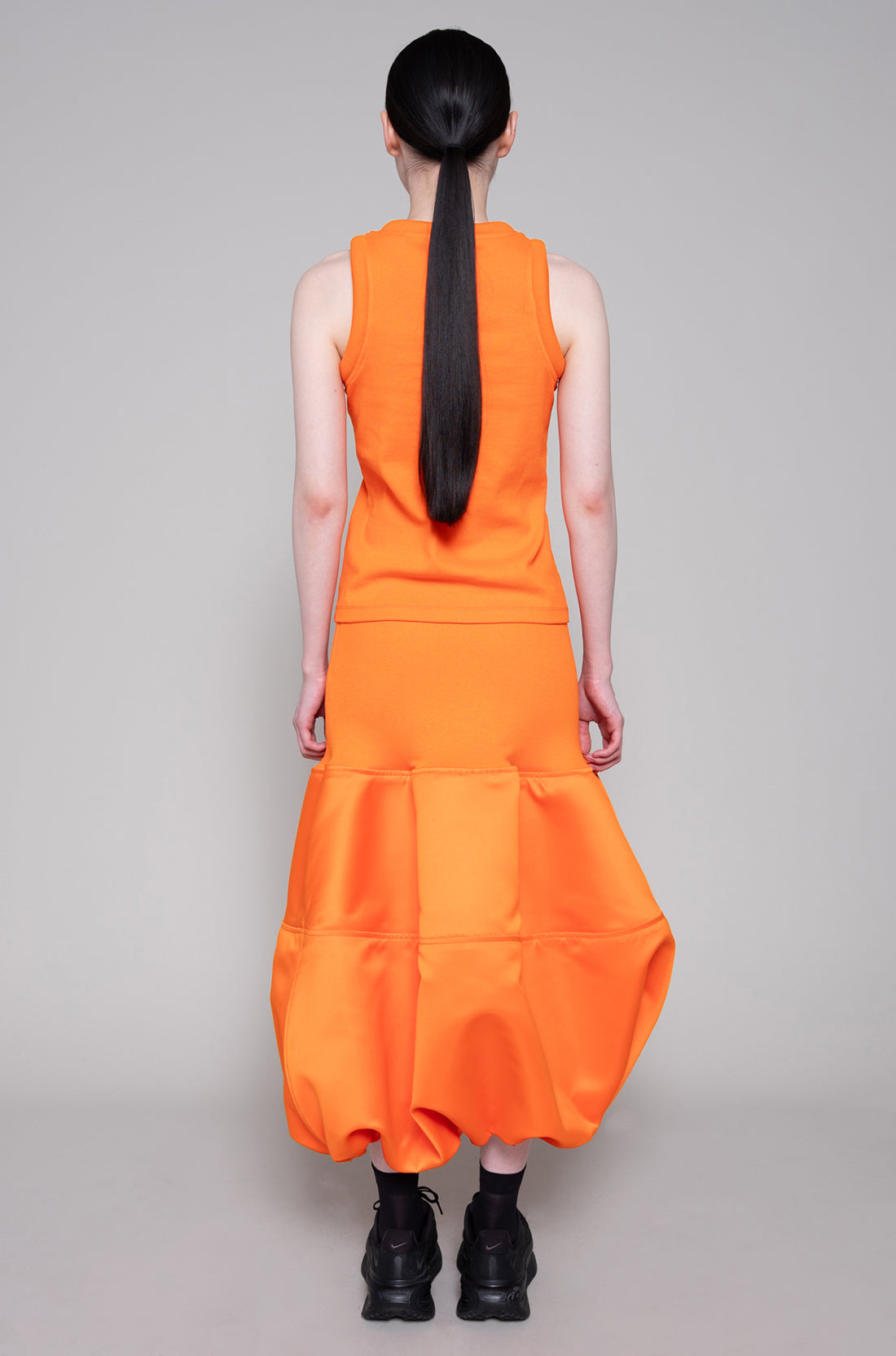 Person wearing sleeveless orange top and skirt with black sneakers on plain background