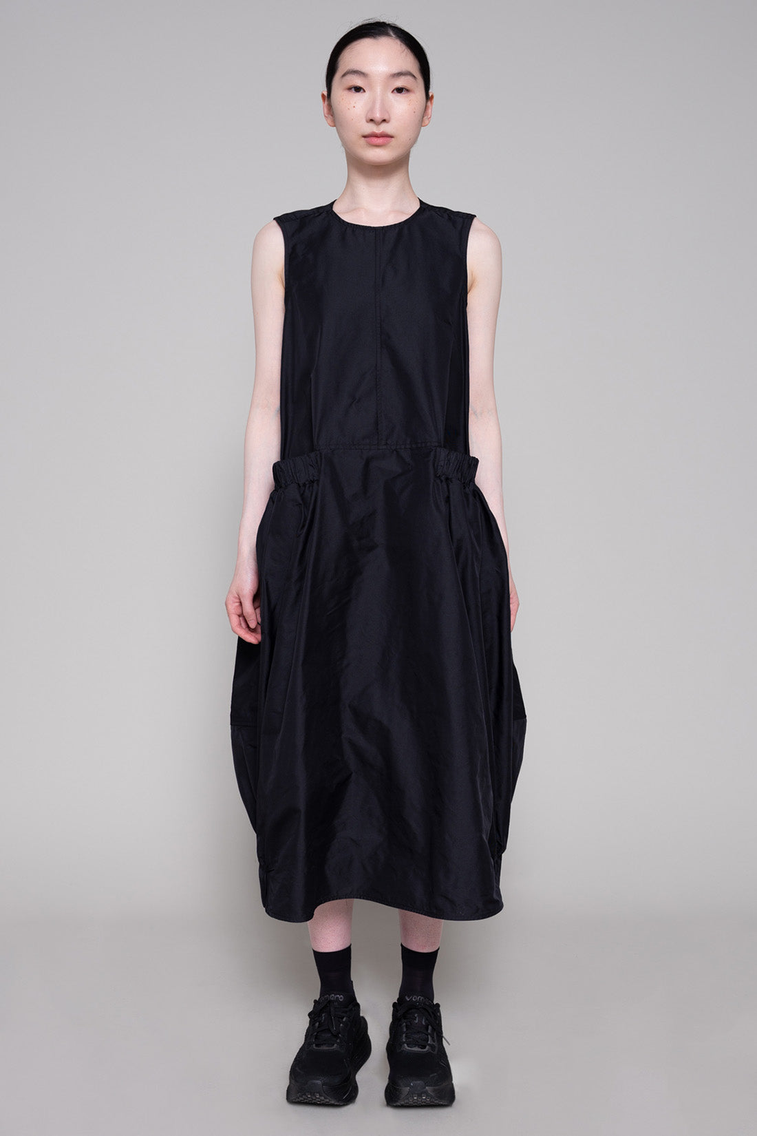 Person wearing a black sleeveless dress and sneakers against a plain background