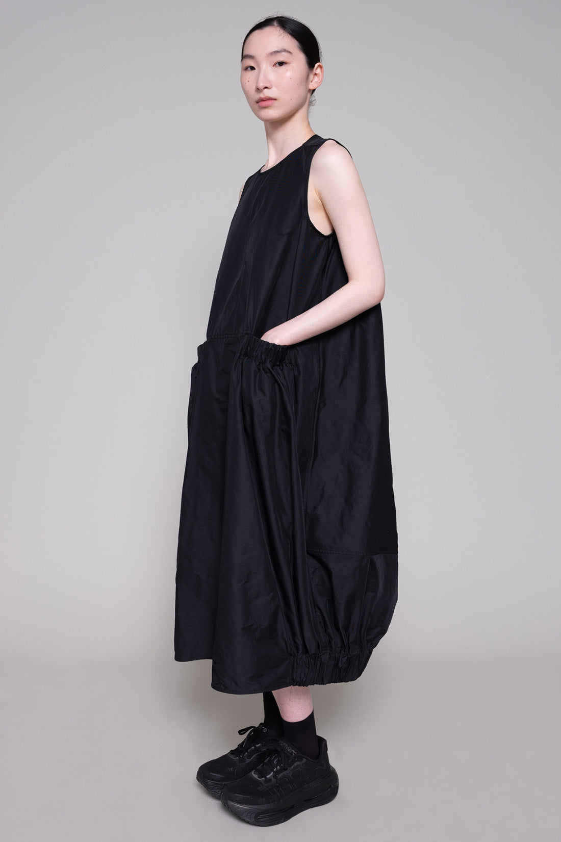 Person wearing a black sleeveless dress and sneakers against a plain background