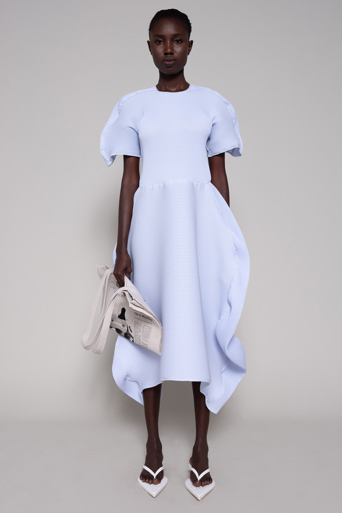 Person wearing a light blue dress and white flip flops holding a tote bag against a plain background