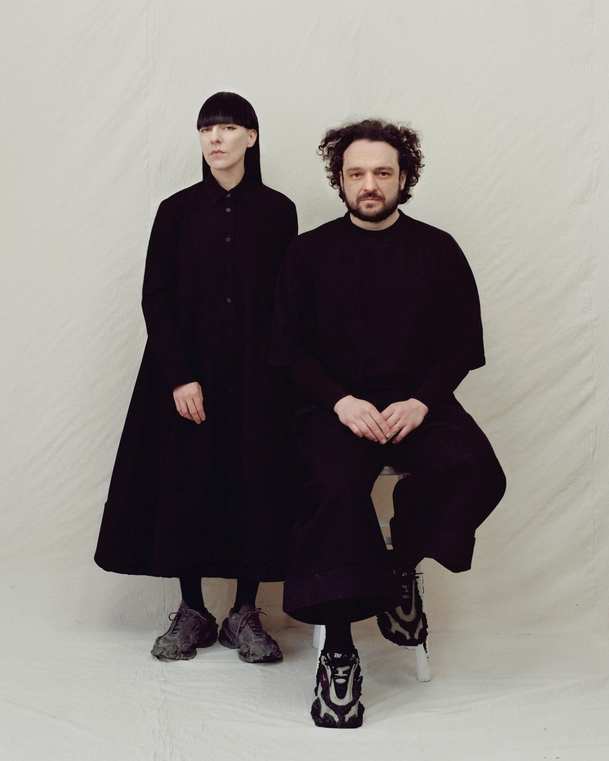 MELITTA BAUMEISTER and MICHAL PLATA wearing oversized black garments, one standing and one seated, against a light off-white fabric backdrop. Photo by Cyril Marion.