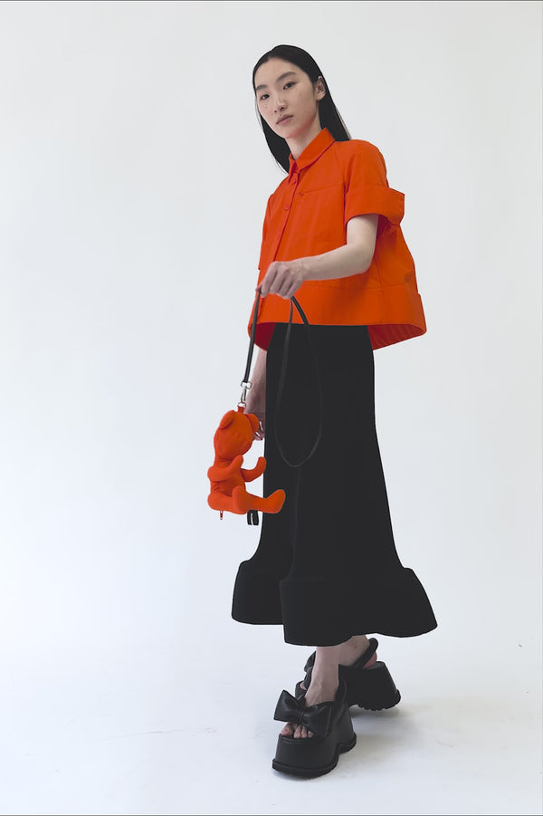 MELITTA-BAUMEISTER-Ripple-Wavy-Skirt-Black-MOVEMENT-VIDEO-MODEL-TURNS-AND-WALKS-FRONT-THEN-OUT-WEARING-ORANGE-TOP-AND-HOLDING-ORANGE-TEDDY-BEAR-BAG