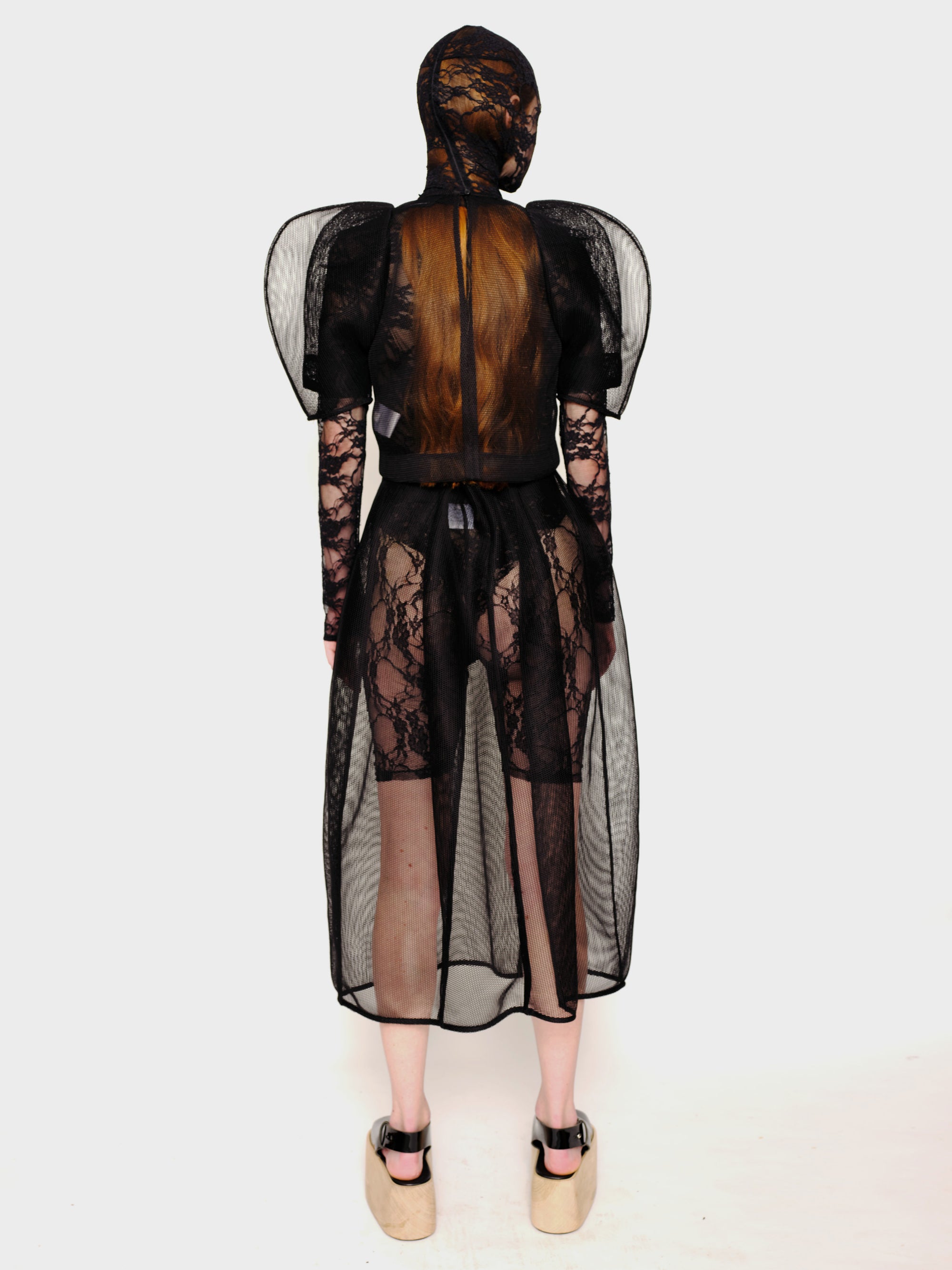 back-angle-of-a-Woman-standing-against-white-background-wearing-Melitta-Baumeister's-black-head-covering-under-layer-covered-by-sheer-lace-dress-with-mesh-fabric-and-puffed-shoulders-and-wearing-platform-sandals