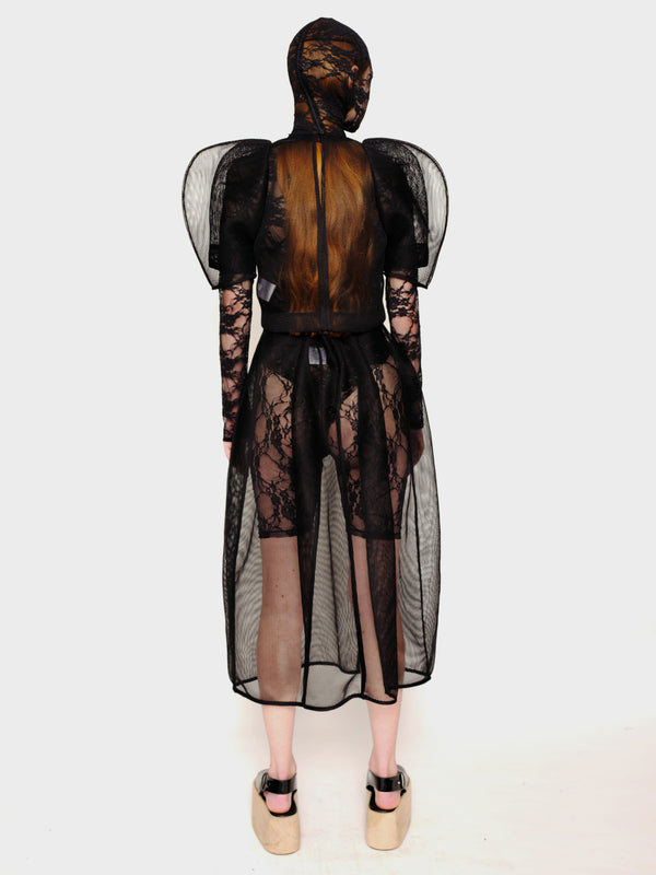 back-angle-of-a-Woman-standing-against-white-background-wearing-Melitta-Baumeister's-black-head-covering-under-layer-covered-by-sheer-lace-dress-with-mesh-fabric-and-puffed-shoulders-and-wearing-platform-sandals