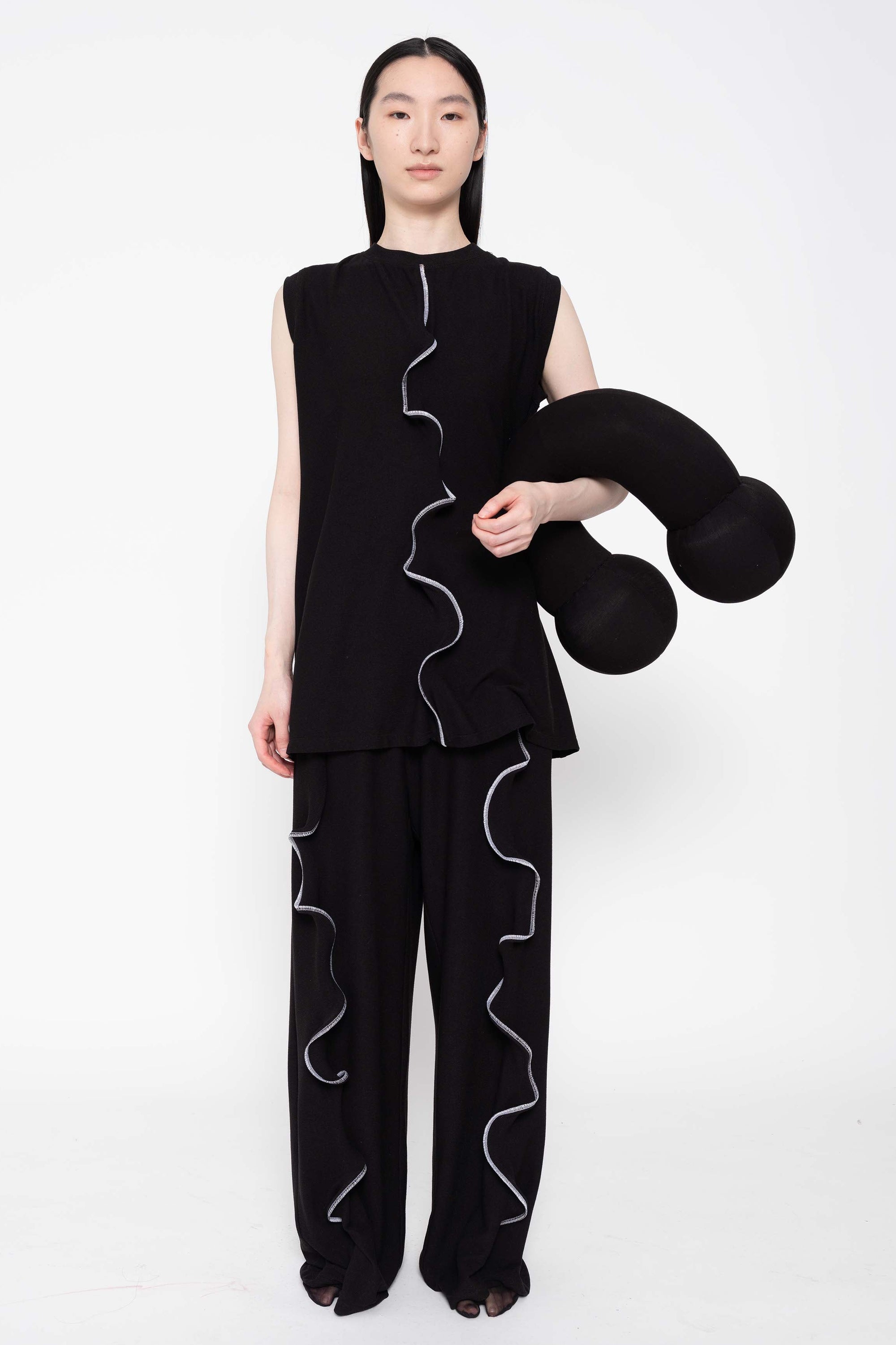 A person standing against a white background, holding a black textured object, wearing black casual pants with a wiggly seam design.