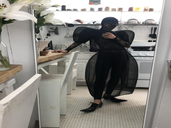 Woman-standing-in-the-kitchen-wearing-Melitta-Baumeister's-black-head-covering-under-layer-covered-by-sheer-lace-dress-with-mesh-fabric-and-puffed-shoulders-paired-with-back-pointy-shoes